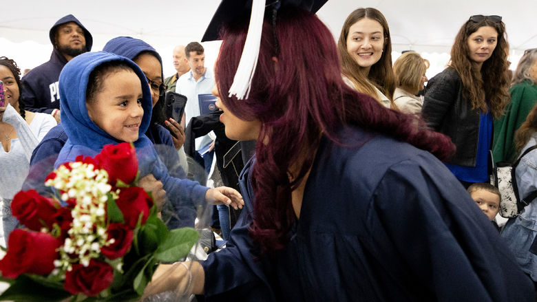 Woman in commencement gown bends down to accept a bouquet of flowers from a young boy.