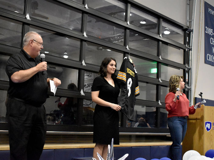 Auctioneers Chuck Johnson, left, and Jodi August, right, gives details on a signed Steeler Kenny Pickett jersey that is being displayed by Anna Raffeinner, center