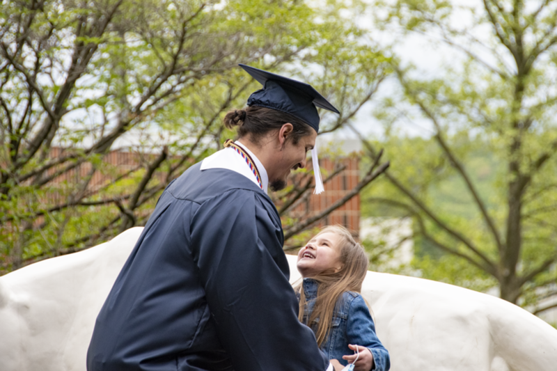 A man in academic regalia including picks up his small daughter in front of Schuylkill's lion shrine.