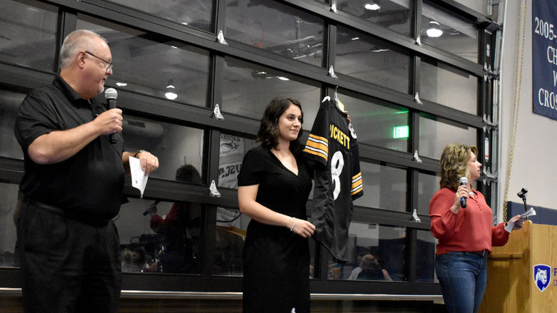 Auctioneers Chuck Johnson, left, and Jodi August, right, gives details on a signed Steeler Kenny Pickett jersey that is being displayed by Anna Raffeinner, center