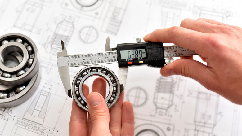 A quality engineer measuring a ball bearing with calipers to ensure its compliance, with a reference diagram in the background.