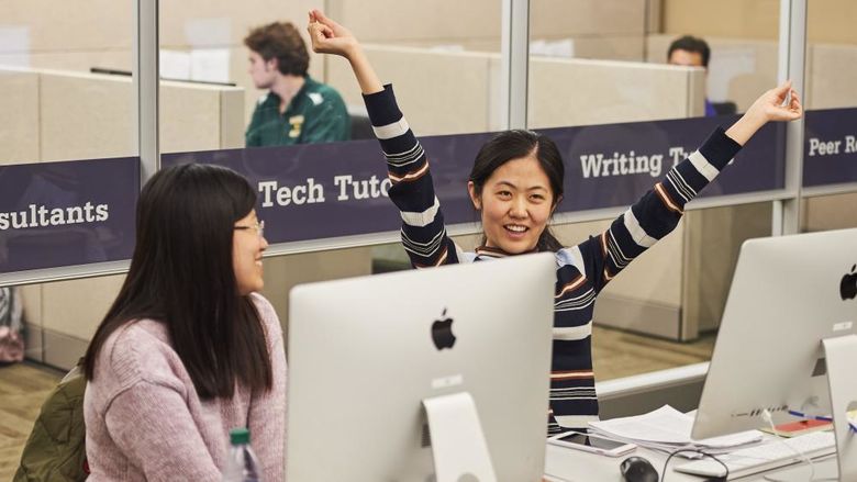 two students sitting together at computer stations, student at right smiles with arms raised joyfully