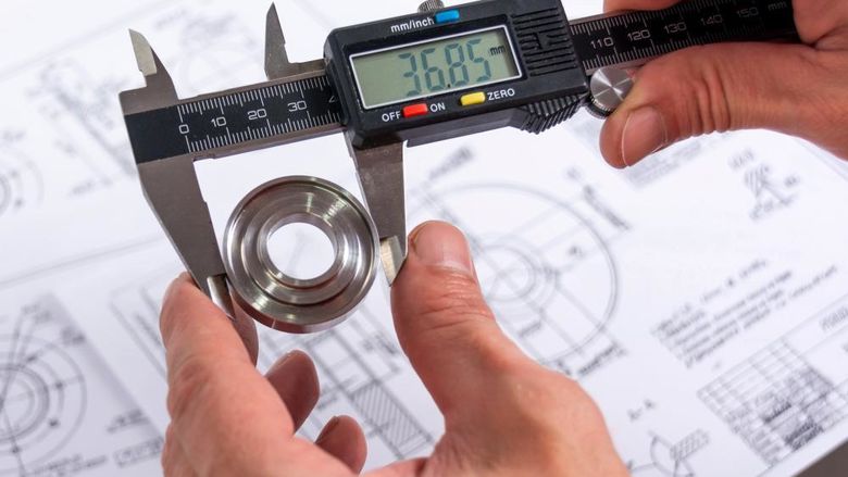 A quality engineer measuring a production part with calipers to ensure its compliance, with a reference diagram in the background.