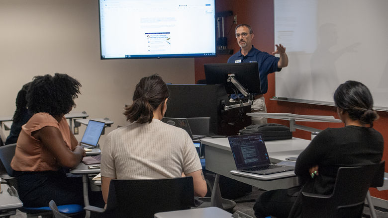 Four people sit in a semicircle in a classroom listening to speaker and watching a screen