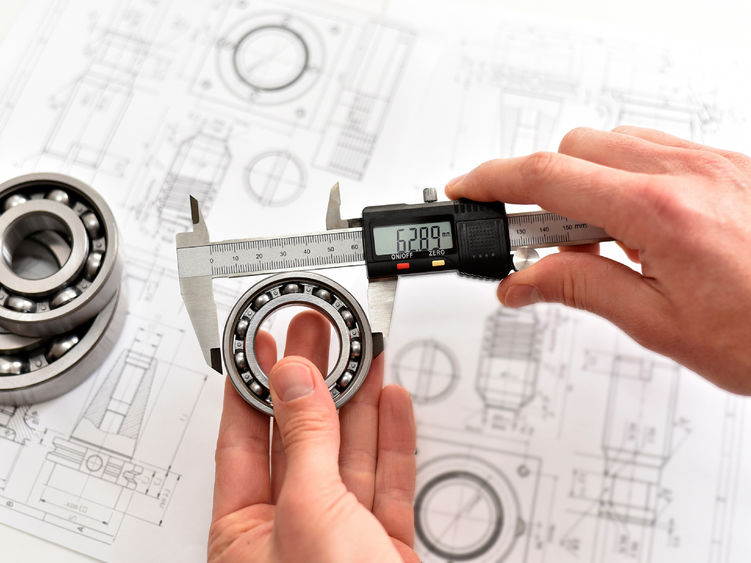A quality engineer measuring a ball bearing with calipers to ensure its compliance, with a reference diagram in the background.