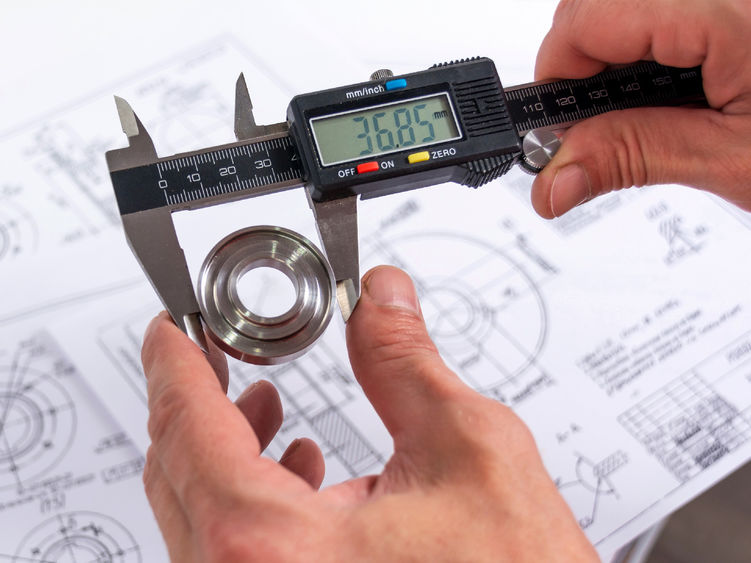 A quality engineer measuring a production part with calipers to ensure its compliance, with a reference diagram in the background.