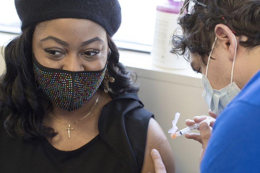 Tamika Washington, who wears a jeweled face mask, a cross necklace and a beret, looks away from her bare arm as a health care worker prepares to insert a syringe filled with the COVID-19 vaccine.